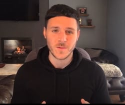 Image shows a still from Ben Andrews YouTube Channel. Here, he is sitting in a living room, wearing a black hooded top.