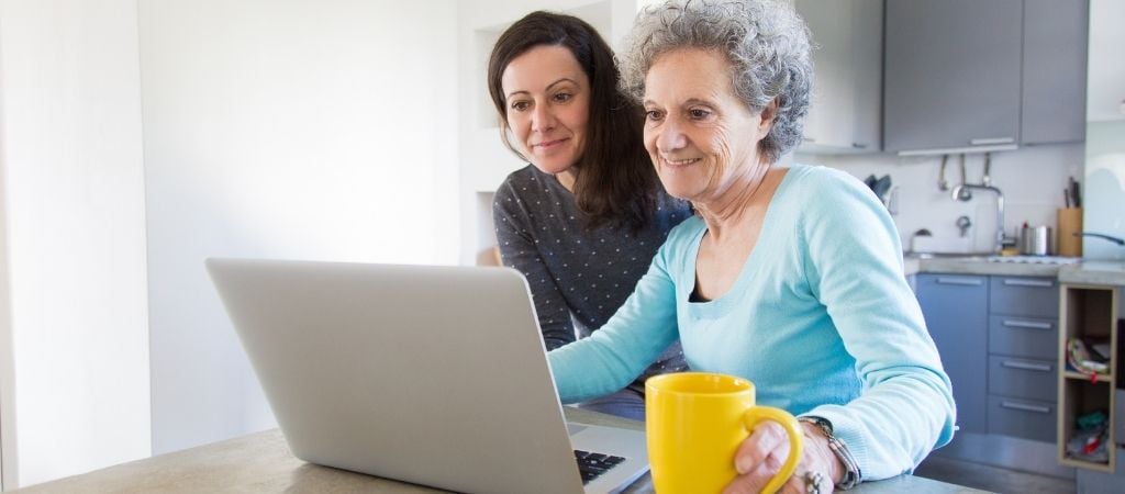 Two ladies looking at a laptop on the kitchen worktop. a yellow mug is next to the open laptop