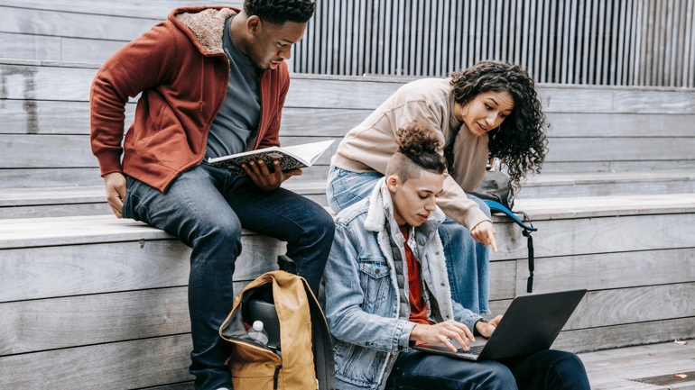 Three students sitting on some steps outdoors, collaborating and looking at a laptop screen on the middle student's lap