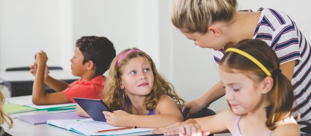 Child holding tablet at school desk in class looks up at her smiling teaching assistant, while other children in the class work