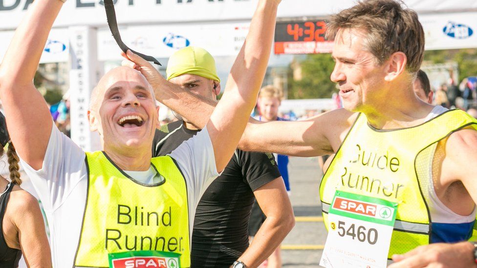 Chris Blackabee smiles and celebrates with his arms in the air at the end of a race, alongside Guide runner Colin Johnson. Both are wearing high vis vests and their race numbers the race finish line can be seen in the background