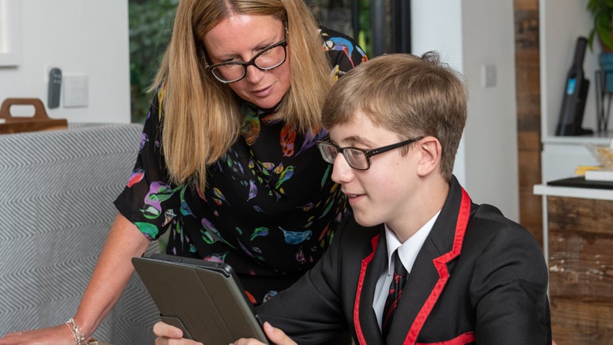 Melanie and Alfie chatting at a desk. Alfie is wearing his school blazer and showing his mum his tablet screen