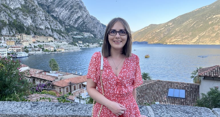 Elin is standing against a low wall, she is wearing a pink and white dress and a small white bag. Behind Elin is a view of a lake and mountains. 