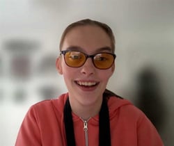 Iona smiles at the camera.  She has dark hair, tied back and wears yellow tinted glasses.  she is also wearing a salmon pink hoody. 