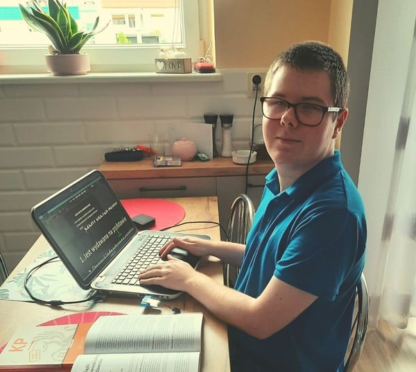 Image shows student Jarosław Hyżyk sitting at his desk in front of a window. He is typing on a laptop while a SuperNova USB is plugged in. The information on the screen is magnified. His textbooks are open on the desk next to his laptop.