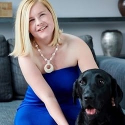 Image shows Sassy Wyatt with her guide dog Ida. Sassy is wearing a blue dress and a white chunky necklace. Sassy is smiling at the cameral and has her arms round Ida, a black lab