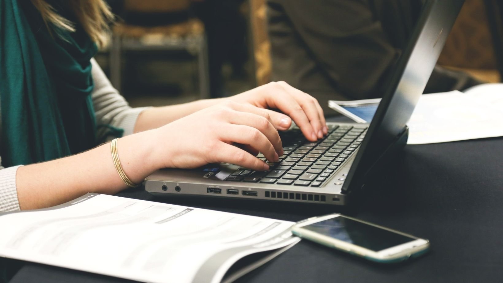 Image description: Woman typing on a laptop computer, with paper notes and smartphone on the table next to her.