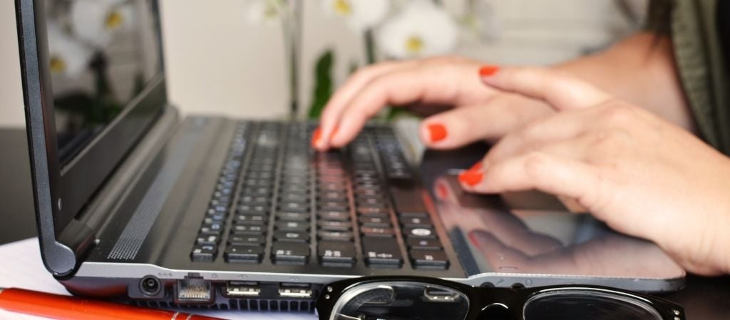 Side view of a laptop. Hands are shown with red nailvarnish typing. a pair of glasses are shown in the foreground.