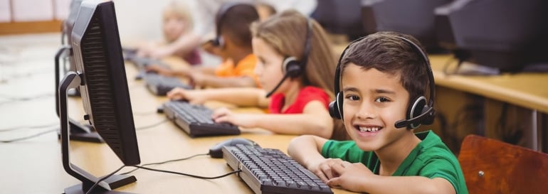 Young students in class on computers. Boy in the foreground is smiling. He is sitting in front of his computer screen and keyboard and is wearing headphones.