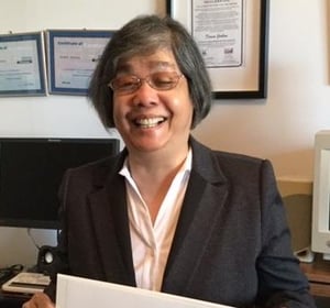 Donna is smiling and holding a certificate as she stands in an office with more certificates framed on the wall behind her