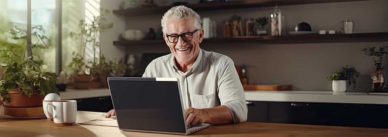 Man with grey hair and glasses is smiling and using a laptop. He is sitting at a counter in a sunny kitchen, with house plants and kitchen jars displayed on shelves behine him